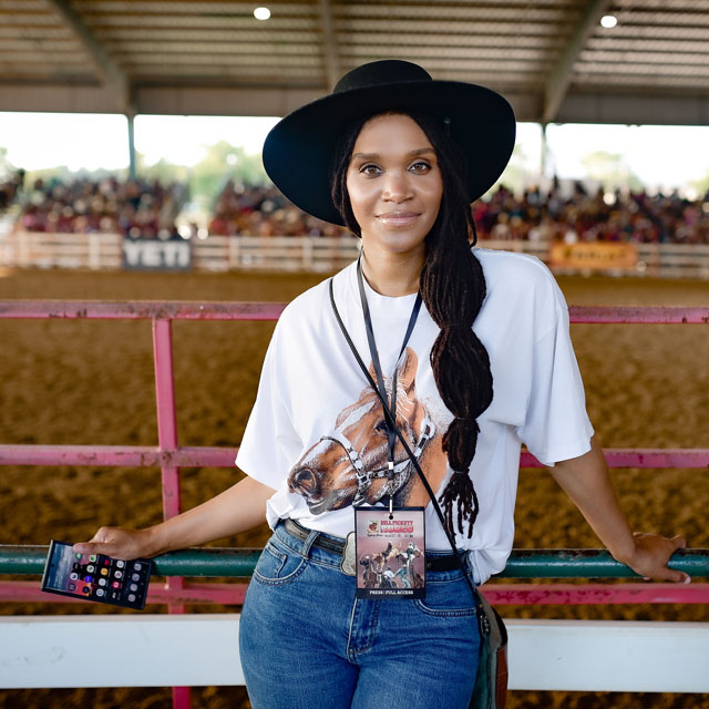 A woman wearing a hat looking directly at the camera, holding a cell phone and posing.